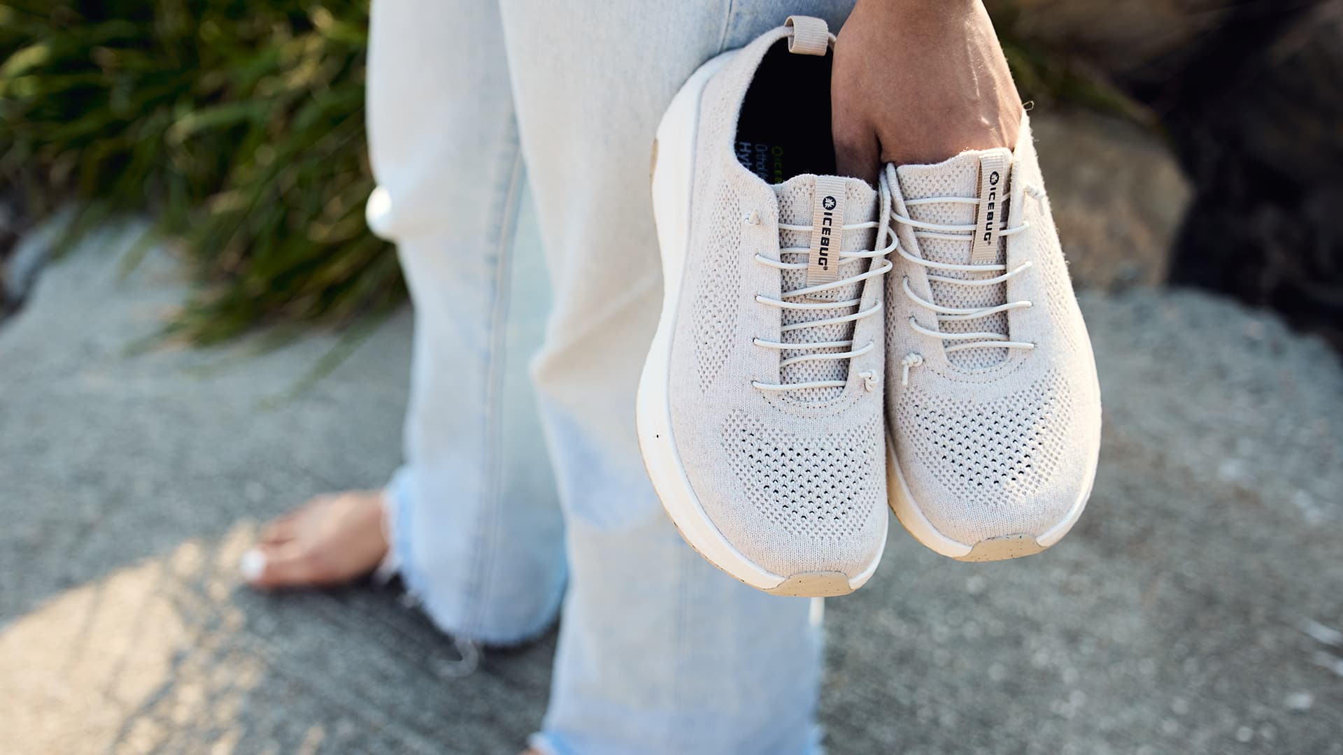 Person holding a pair of light gray sneakers with white laces, standing on a concrete surface, wearing light blue jeans.